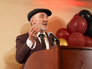 An elderly person in a dark suit and cap speaks into a microphone at a wooden podium, gesturing with their hand, with red and black balloons in the background.