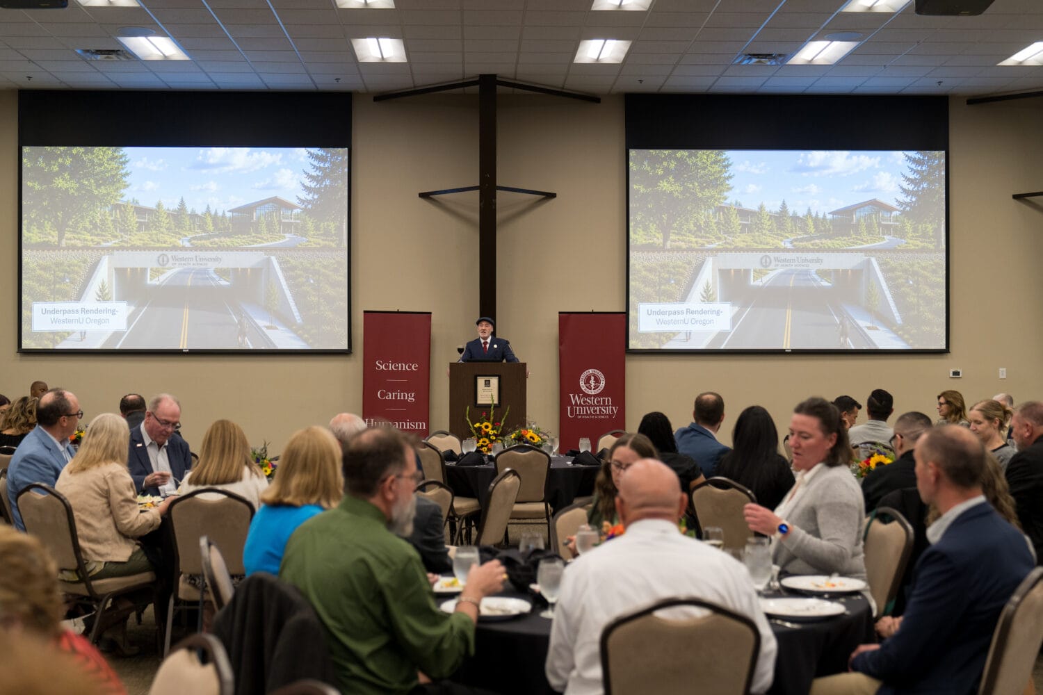 A speaker addresses an audience at a formal event, with large screens displaying an underpass rendering for Western University and banners featuring its mission statement.
