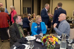 A group of adults engage in conversation at a formal dinner event, seated around a table adorned with a colorful floral centerpiece.