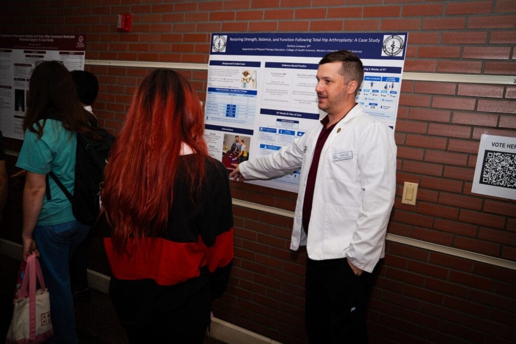 A person in a white lab coat gestures toward a research poster while two others observe it in a hallway with brick walls.