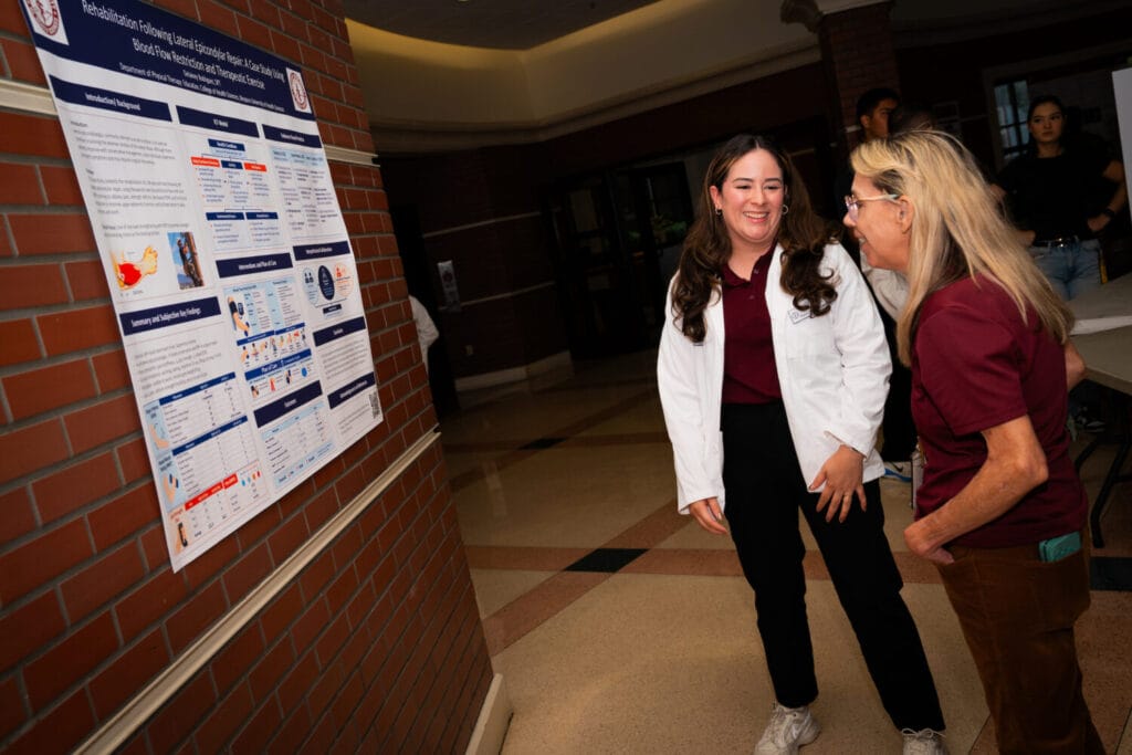 Two individuals engage in conversation near a research poster displayed on a brick wall in an indoor hallway.