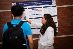 A person in a white lab coat points to a research poster while another individual in a blue shirt observes.