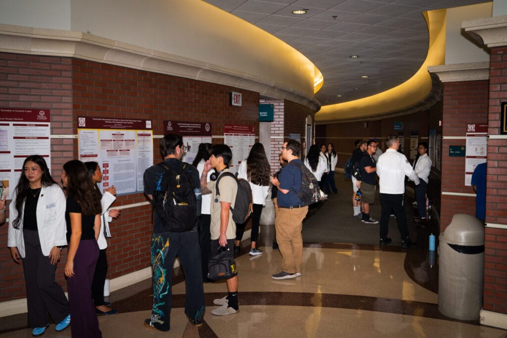 A group of individuals in a hallway with brick walls and illuminated signage, some wearing lab coats and carrying backpacks, are gathered around informational posters.
