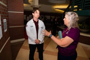A person in a white lab coat stands facing another individual in a purple shirt who is gesturing while speaking, in an indoor hallway with brick walls and a staircase.