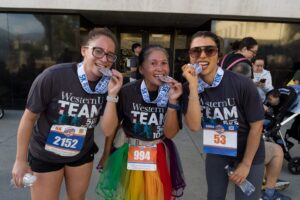 Three individuals wearing "WesternU TEAM" shirts and race bibs smile while holding medals, celebrating their participation in a running event.