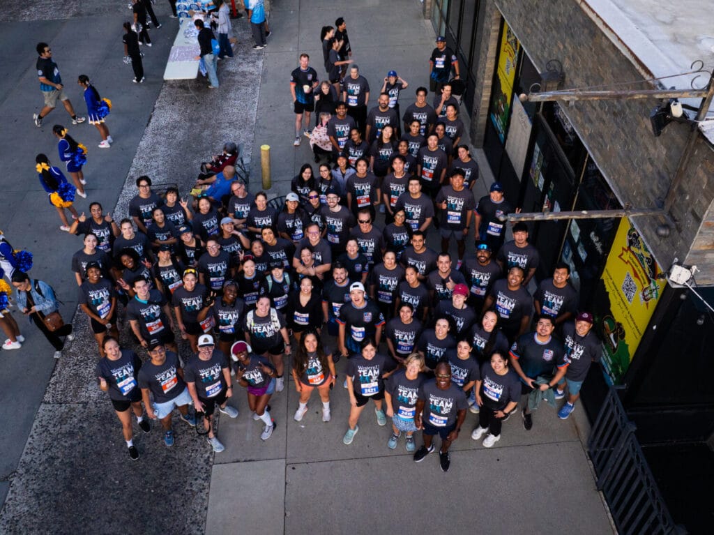 A large group of people wearing matching shirts gather outdoors for a team photo, with cheerleaders and event staff nearby.