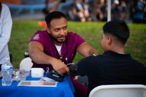 A healthcare worker in maroon scrubs takes a blood pressure reading from a person seated at an outdoor table.