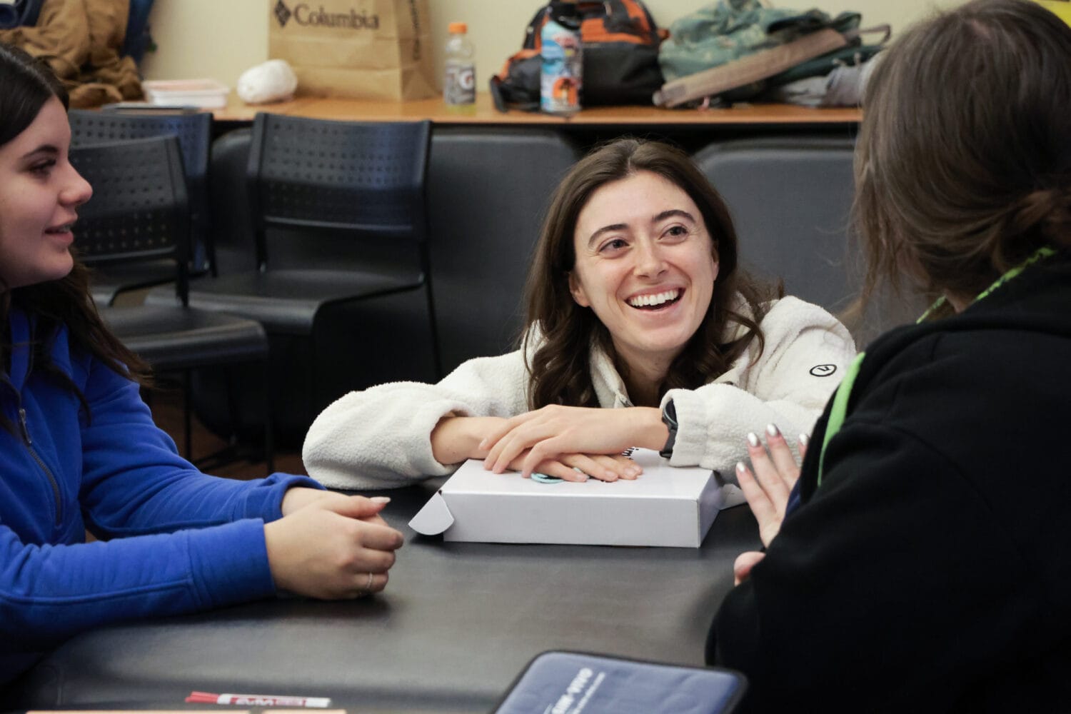 Three young women sit around a table in a classroom, talking and laughing.