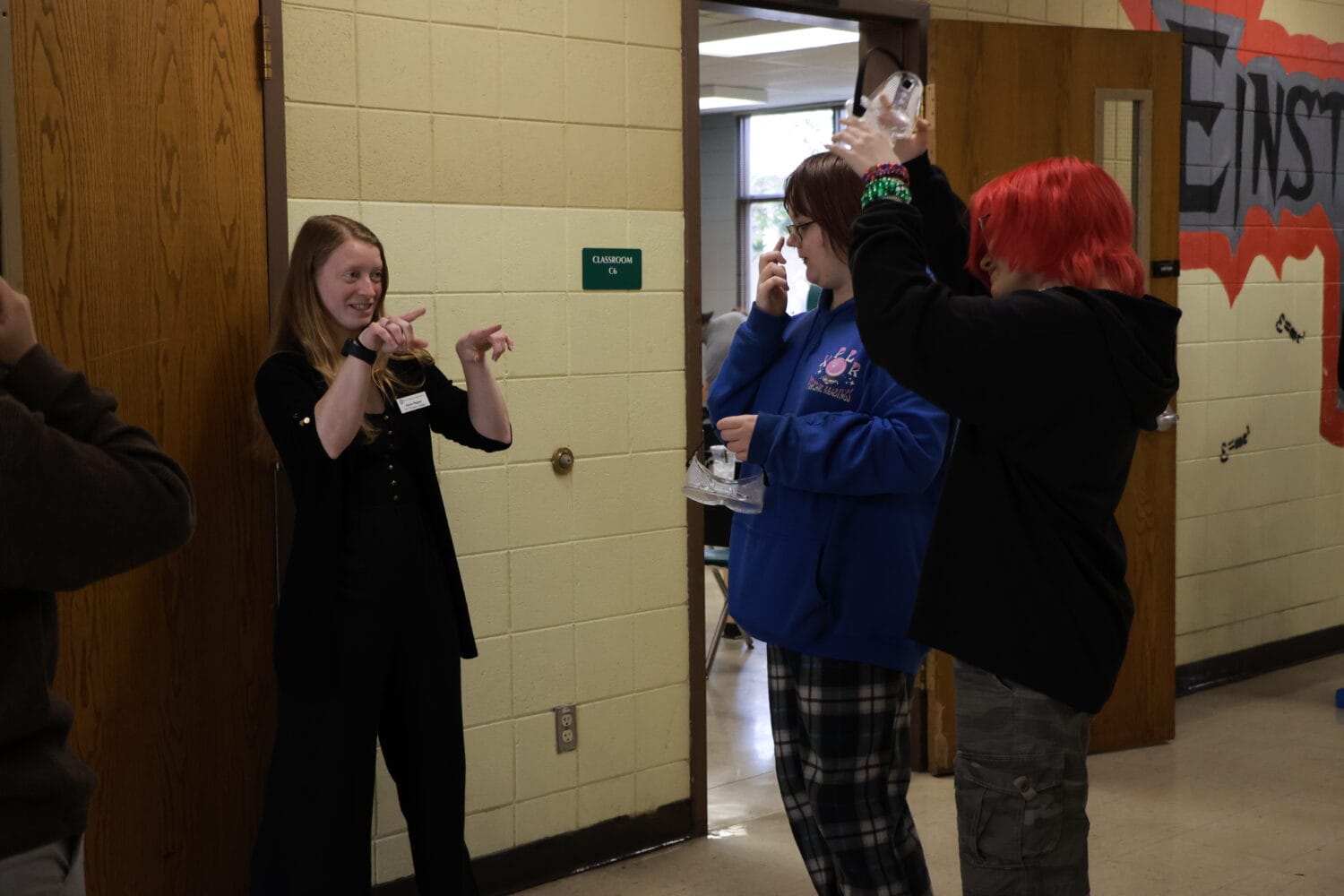 A teacher signs to two students who are holding safety goggles in a school hallway.