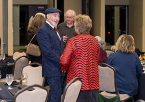 A man in a pinstripe suit and cap converses with a person in a red patterned jacket at a formally set dining event.