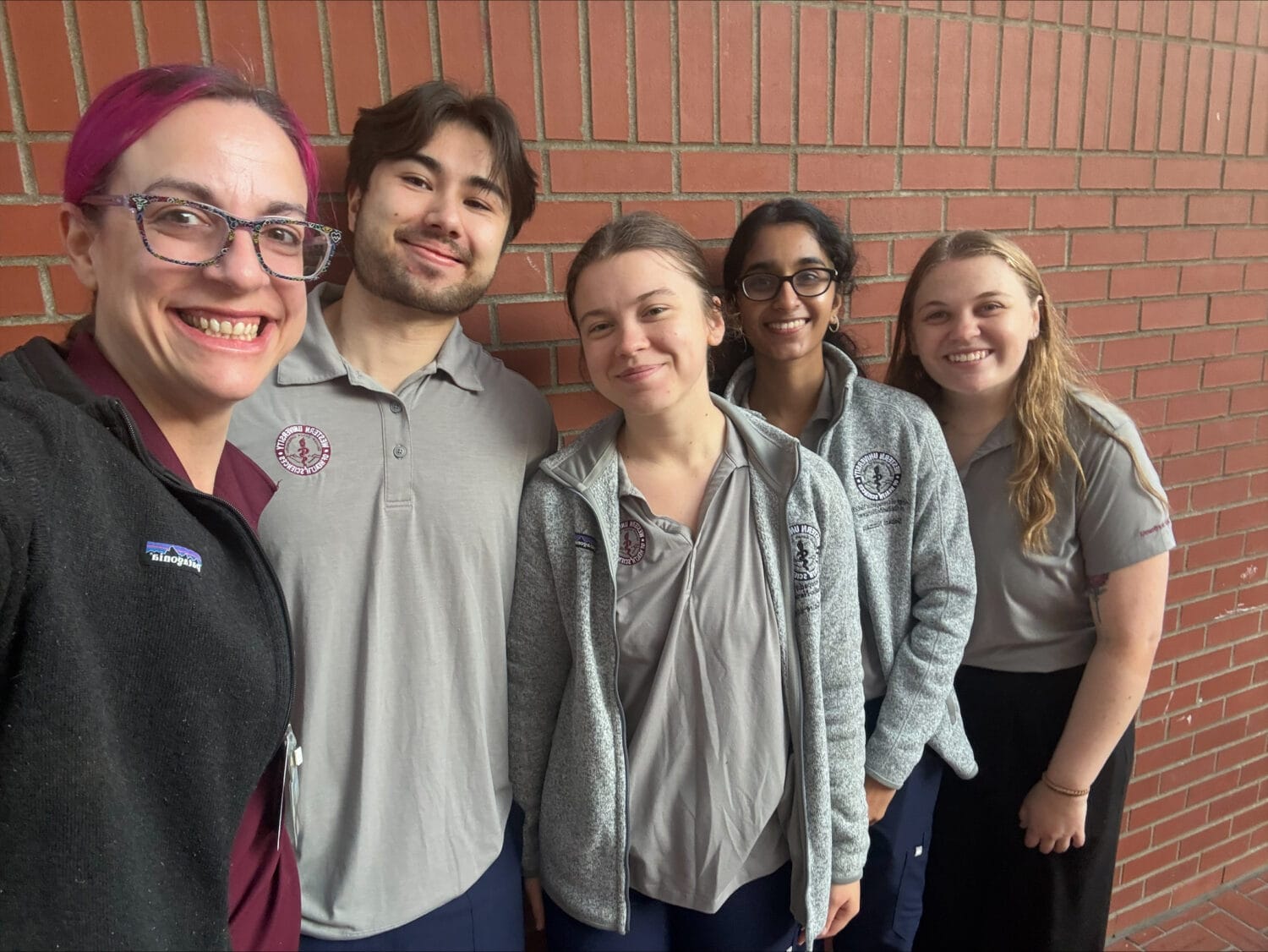 Five people in grey shirts and jackets standing in front of a brick wall.