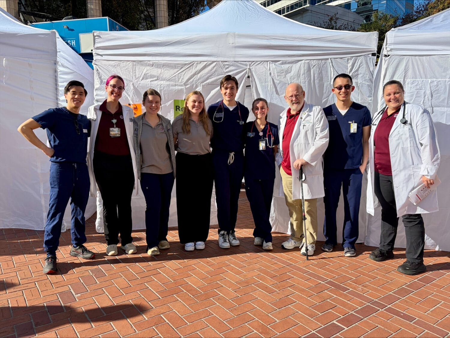 A group of eight healthcare workers in scrubs and lab coats standing together in front of white vendor tents.