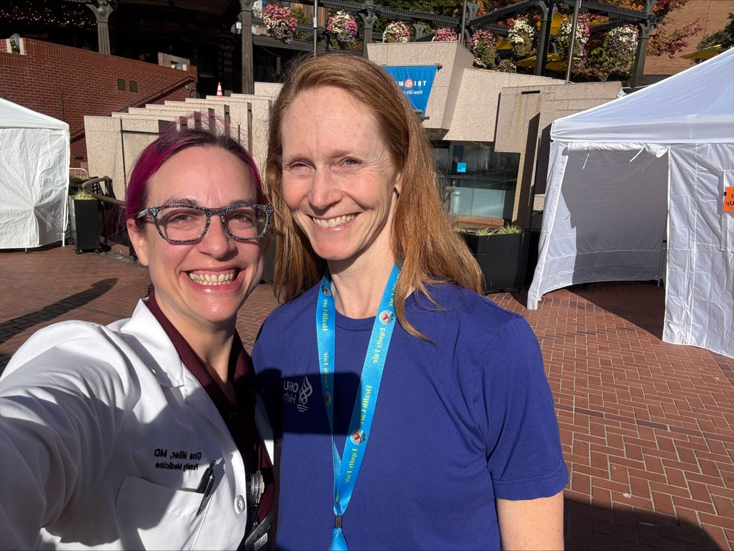 Two women smiling outdoors, one wearing a white doctor’s coat and the other wearing a blue T-shirt with a lanyard.